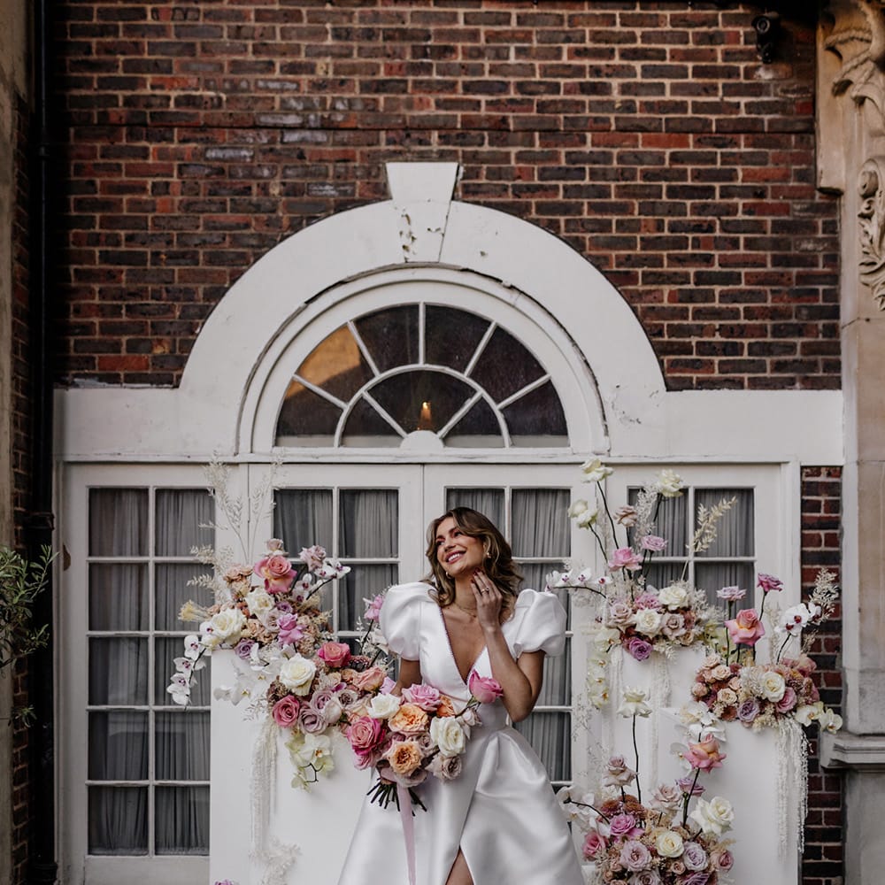 Beautiful bride in a stylish wedding dress with front split holding a pink peeled back rose bouquet standing in front of blush floral arrangements 