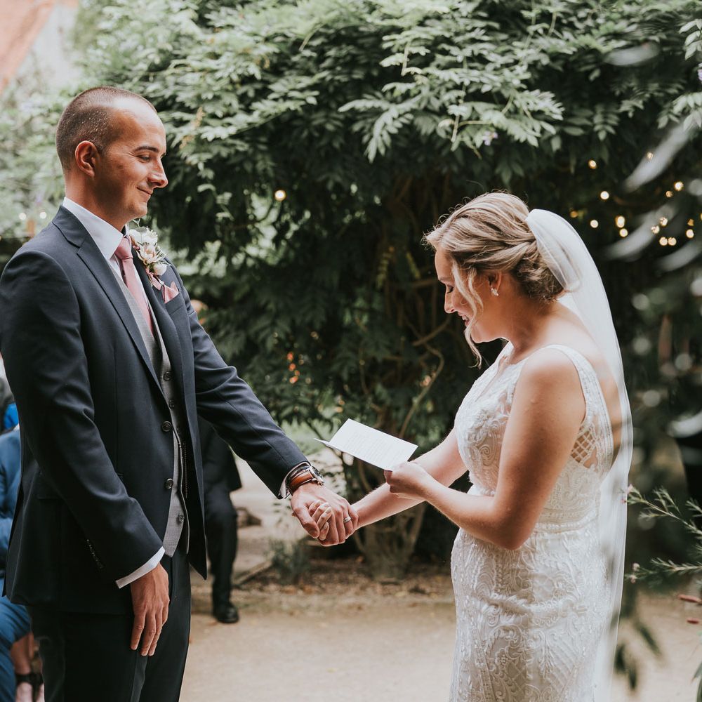 Groom in dark three piece suit with grey waistcoat and pink tie holds hands with bride in lace Justin Alexander wedding dress and veil as she reads vows during outdoor wedding ceremony at Tythe Barn