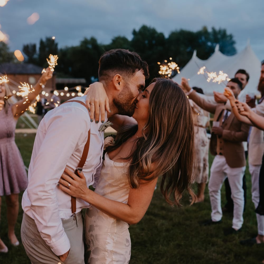 Bride & groom kiss as wedding guests hold sparklers around them for nighttime reception | Mark Bamforth Photography