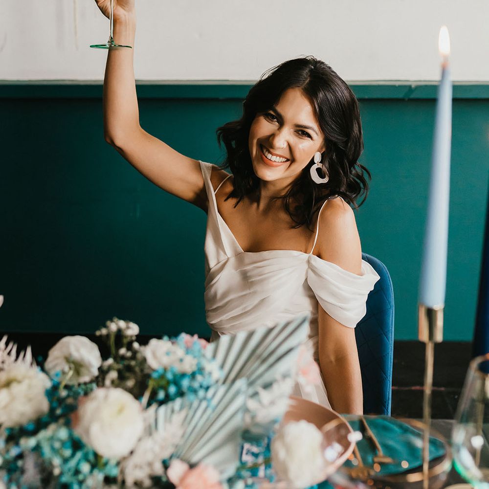 Bride with short wavy hair in a cold shoulder wedding dress holding her coloured glass in the air at her teal wedding 