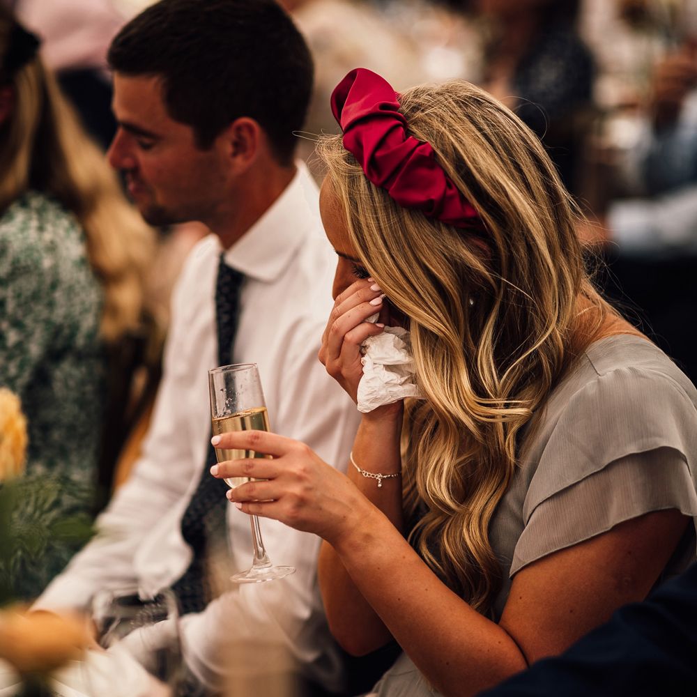 Bridesmaid in grey ruffled bridesmaid dress and pink headband wipes eyes with tissue as she holds glass of Prosecco at garden wedding reception in Cornwall