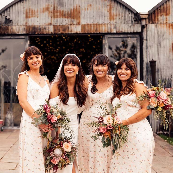 Bridesmaids in cream floral sating bridesmaid dresses posing with the bride holding pink protea wedding bouquets 