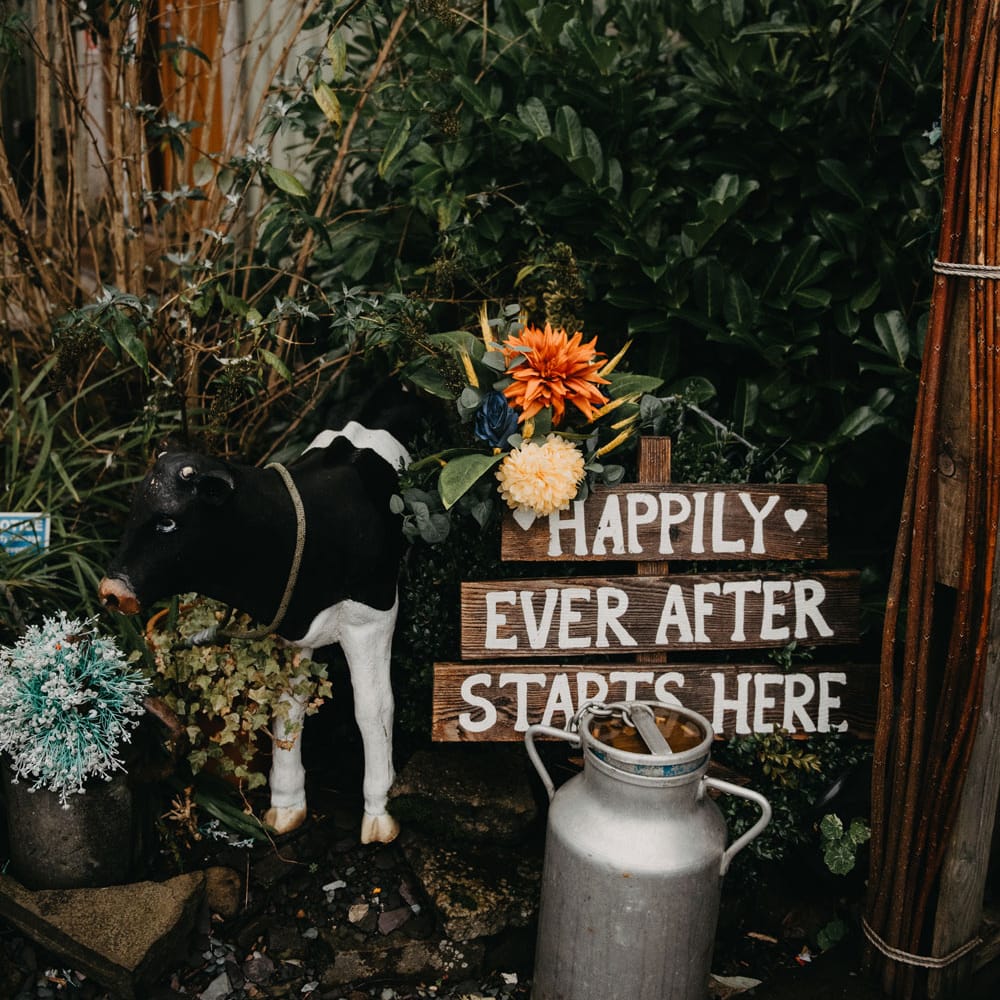 Large floral bouquet by wooden sign reading 'Happily Ever After Starts Here' and milk churn at The Wellbeing Farm for wedding