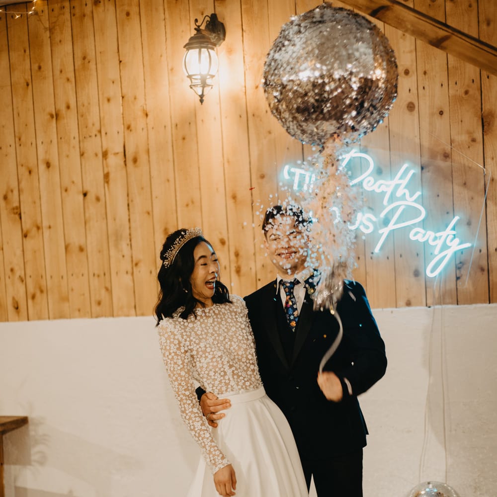 Groom in dark blue suit with floral tie stands with bride in long sleeved applique wedding dress and headpiece as they pull disco ball piñata at rustic wedding ceremony
