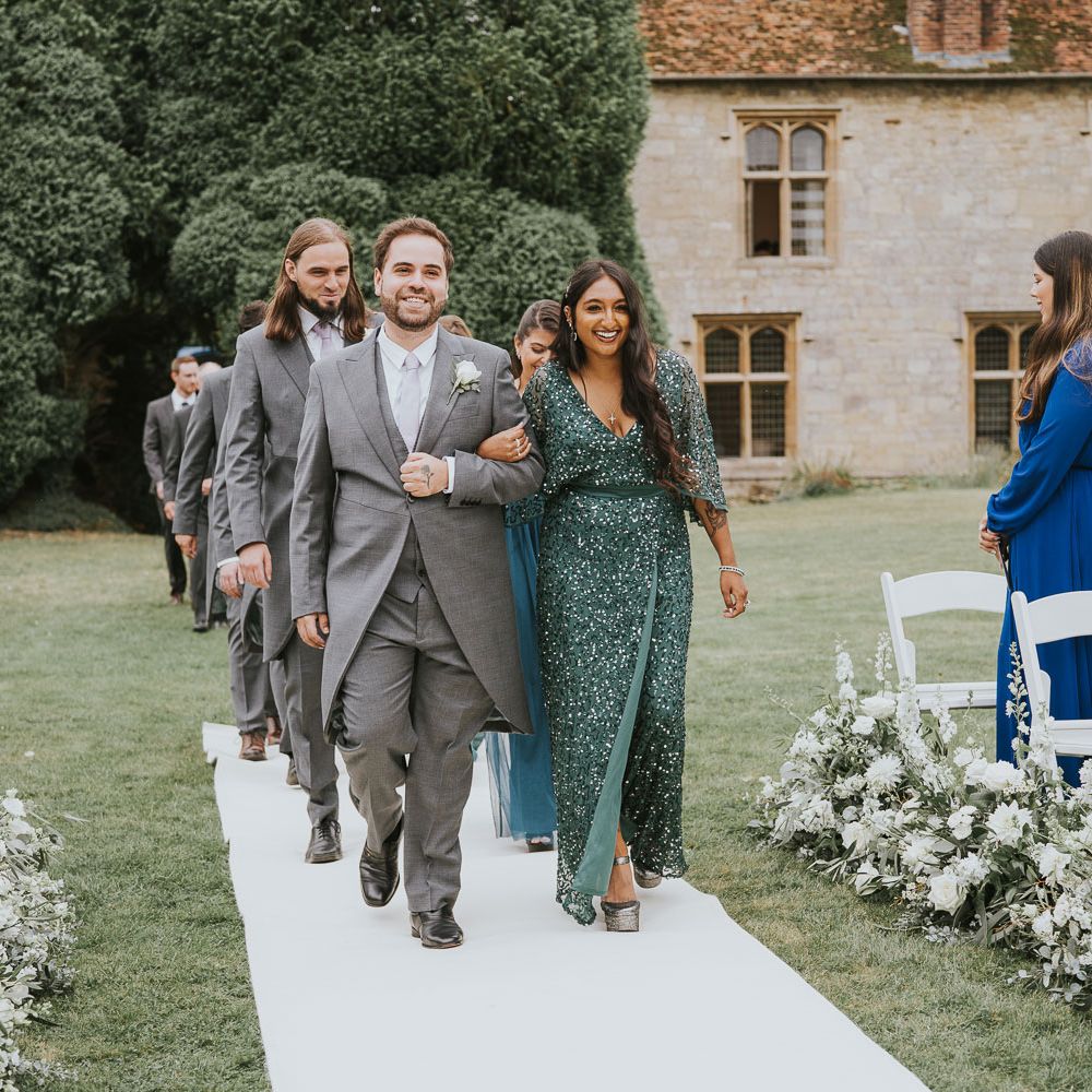 Groomsmen in morning suits walk up the white carpeted aisle with bridesmaids in green bridesmaid dresses during outdoor wedding ceremony at Notley Abbey