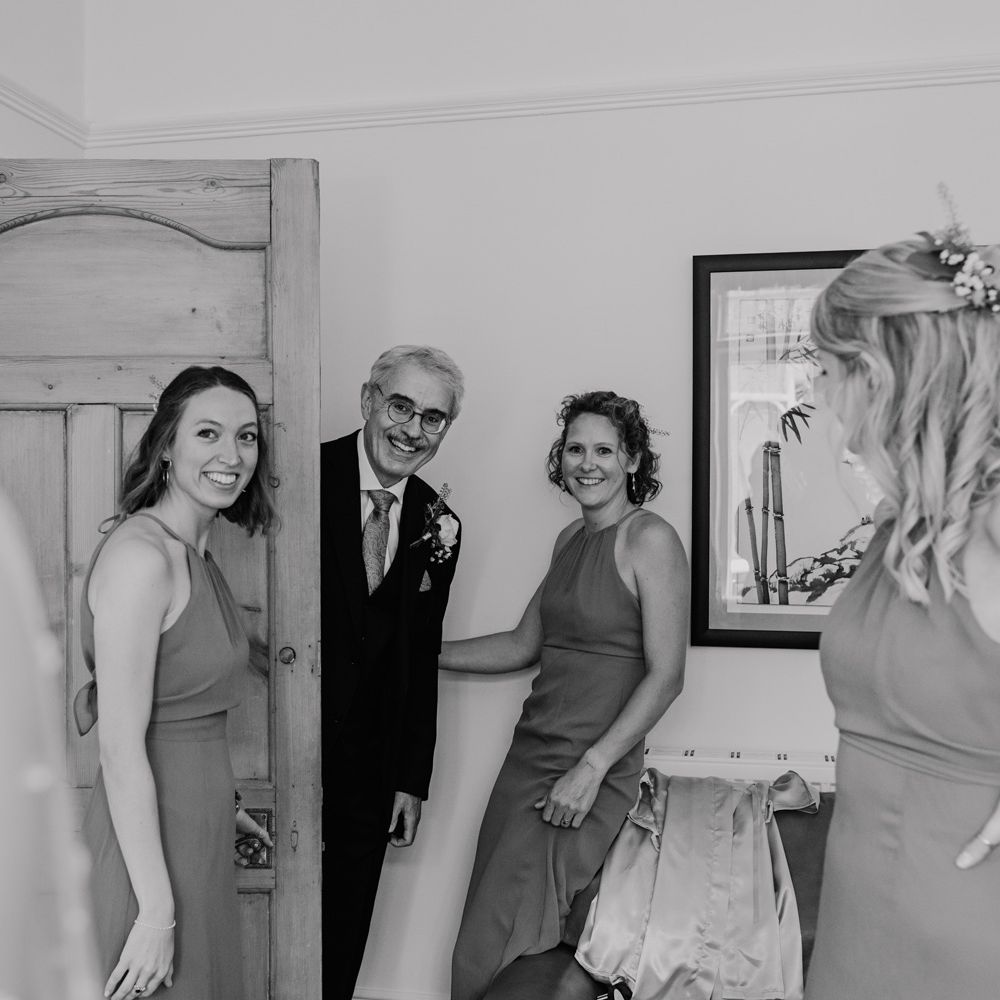 Father of the bride in dark suit and buttonhole smiles as he sees bride for the first time before church wedding ceremony