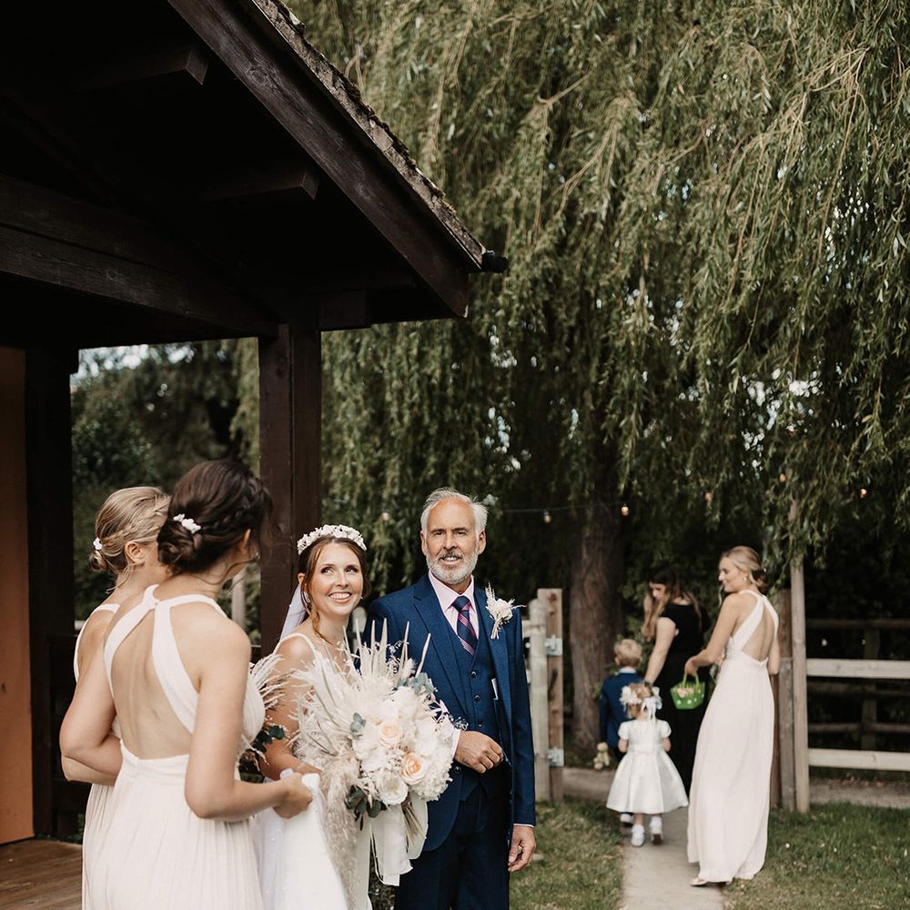 Bride holds her fathers arm and looks toward her bridesmaid outdoors on her wedding day