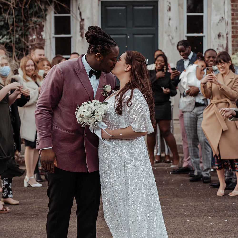 Bride & groom leave ceremony as wedding guests throw confetti around them and they kiss 