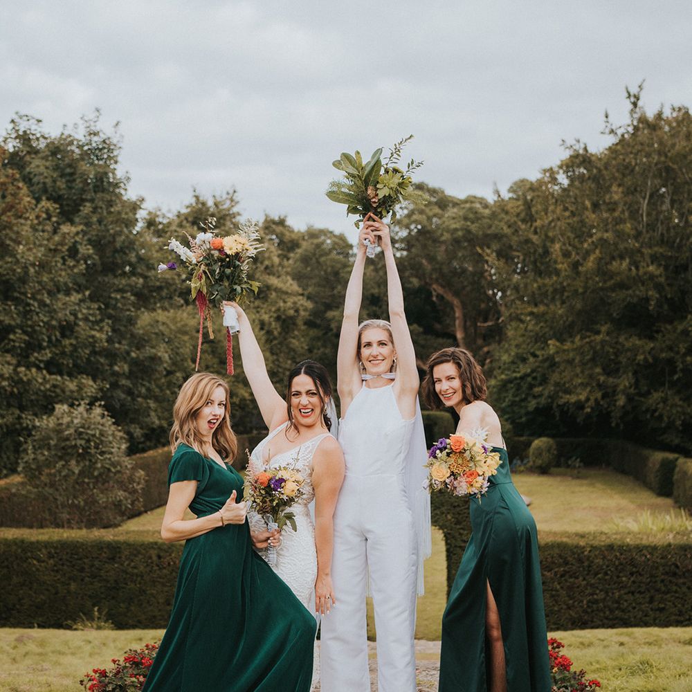 Two brides in a bridal jumpsuit and lace wedding dress with their bridal party in green bridesmaid dresses 
