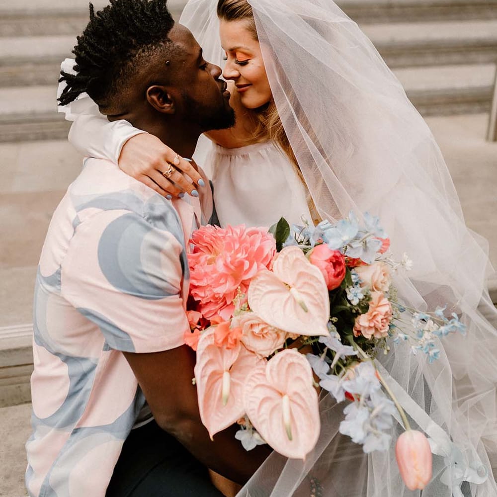 Bride with a long veil and holding a pink and blue wedding bouquet sitting on her grooms lap in a patterned shirt 