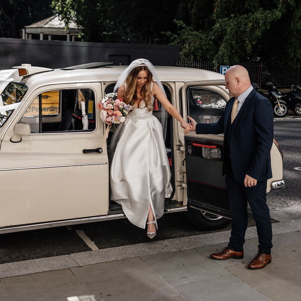 Bride in a Karen Willis Holmes princess wedding dress holding a colourful pink bridal bouquet getting out of a cream London cab 