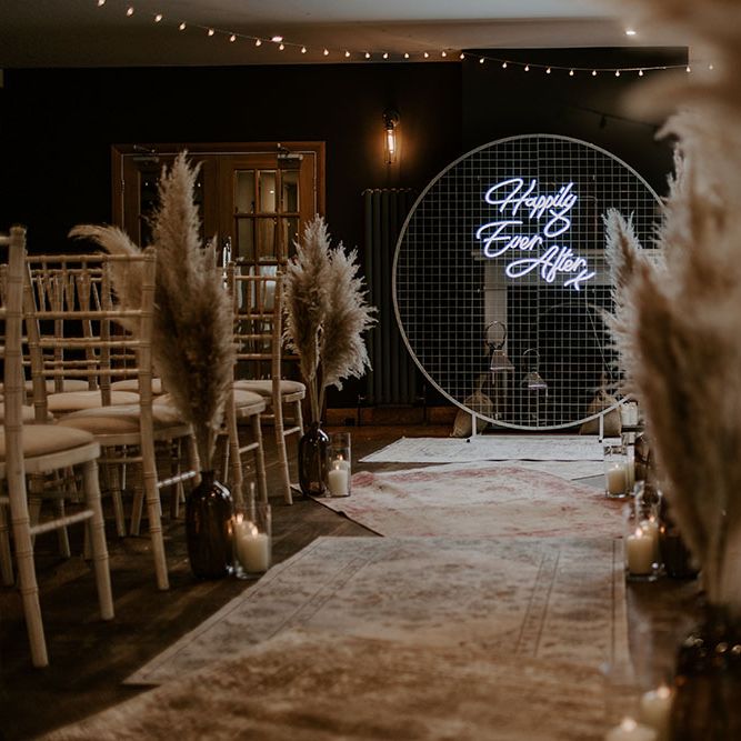 Pampas grass lines aisle alongside white chairs complete with neon sign and Moroccan styled rugs 