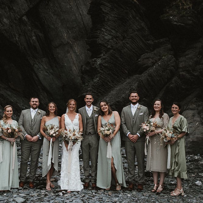 Bride & groom stand with their bridesmaids who wearing different styled green bridesmaid dresses and grooms in three piece suits at Tunnels Beach 