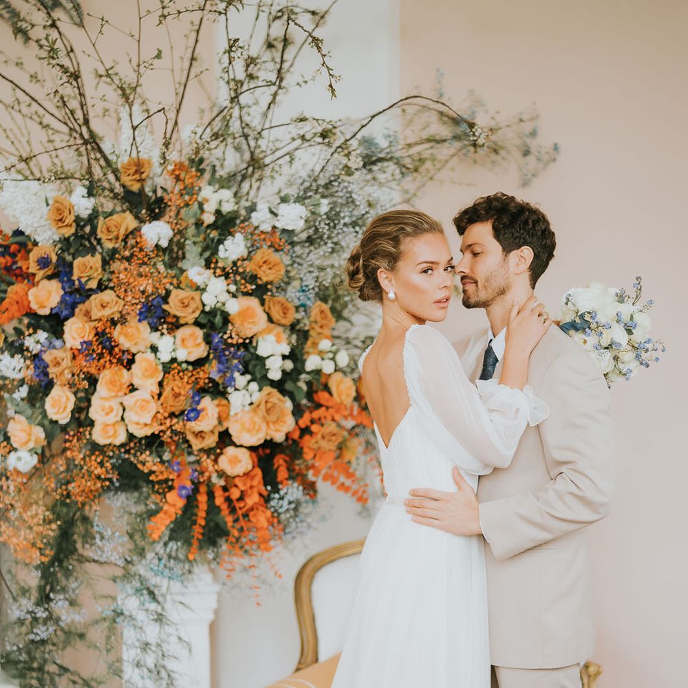 Soft neutral wedding outfits for the bride and groom with the orange and blue wedding flowers in the back 