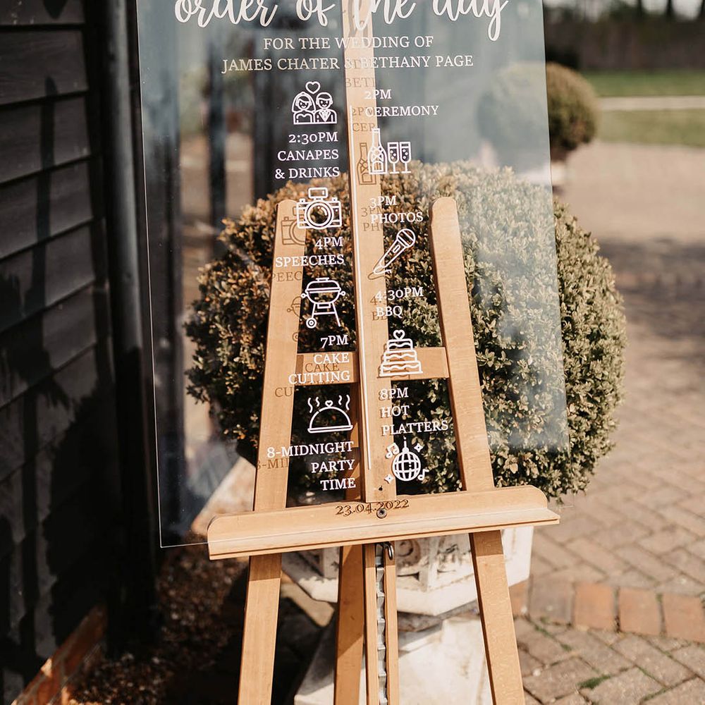 Clear acrylic order of the day wedding sign on a wooden easel with foliage decoration 
