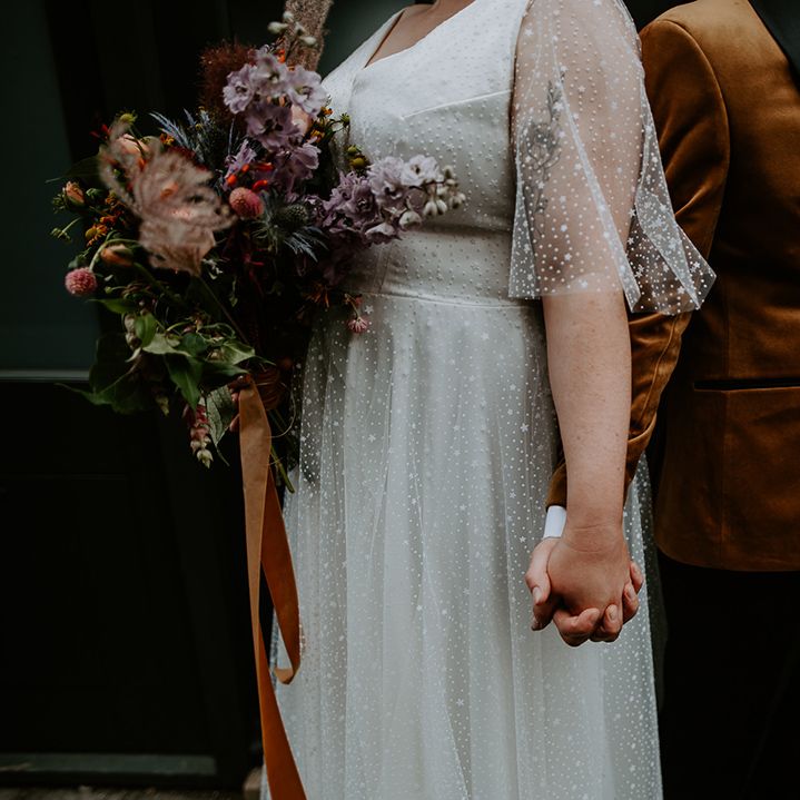 The couple holding hands whilst showing off the bridal bouquet which is tied with a burnt orange ribbon and features Thistles, dried grass and lilac Delphiniums