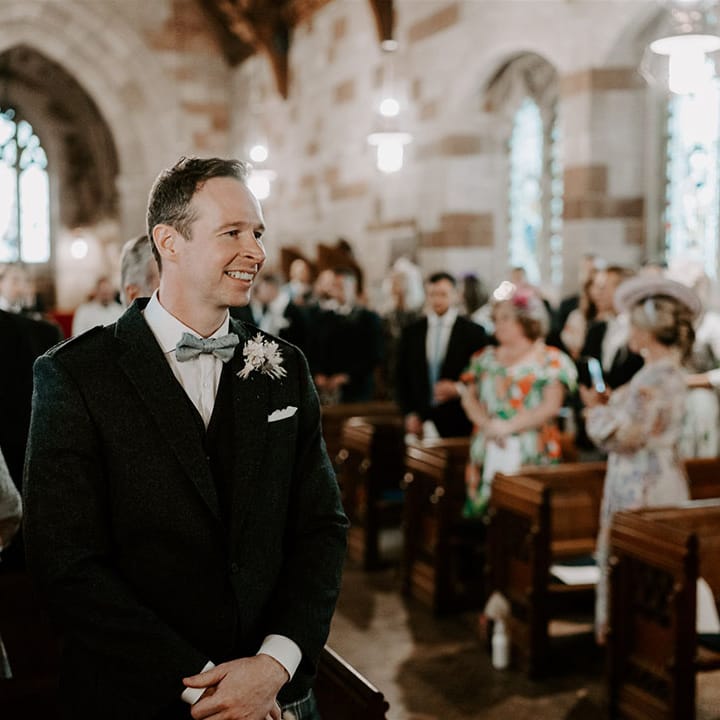 Groom smiling in suit and bow tie with white buttonhole and kilt as he waits for the bride for their church ceremony 