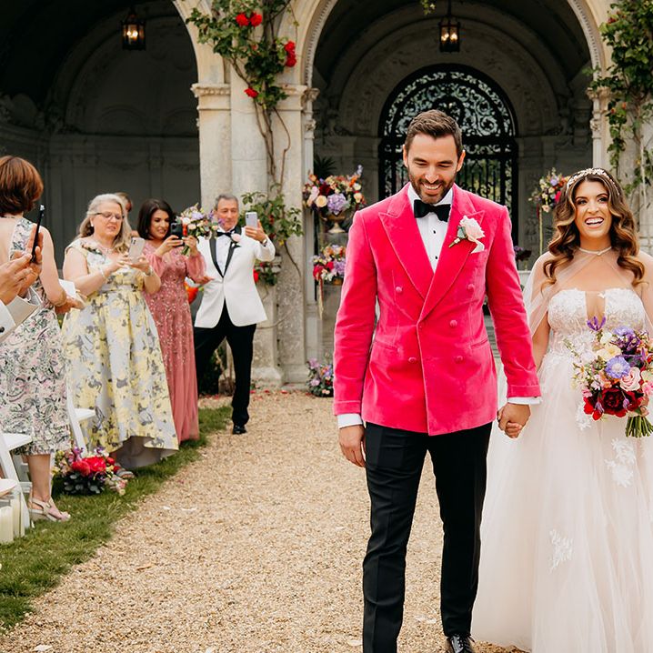 Groom in black bow tie and bright pink velvet suit jacket walking back down the aisle with the bride in a strapless wedding dress with a sheer bridal cape
