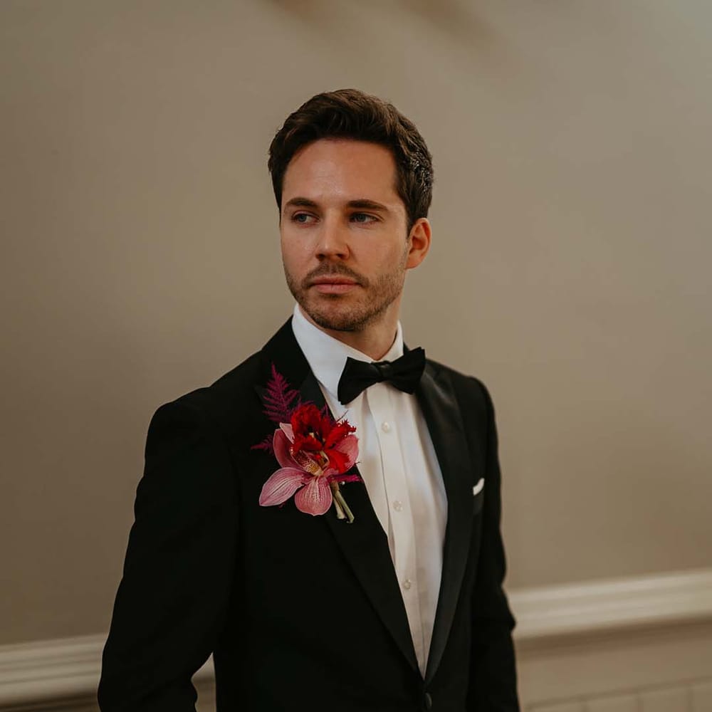 Groom standing and posing wearing classic black tuxedo with magenta amaryllis and orchid boutonniere 