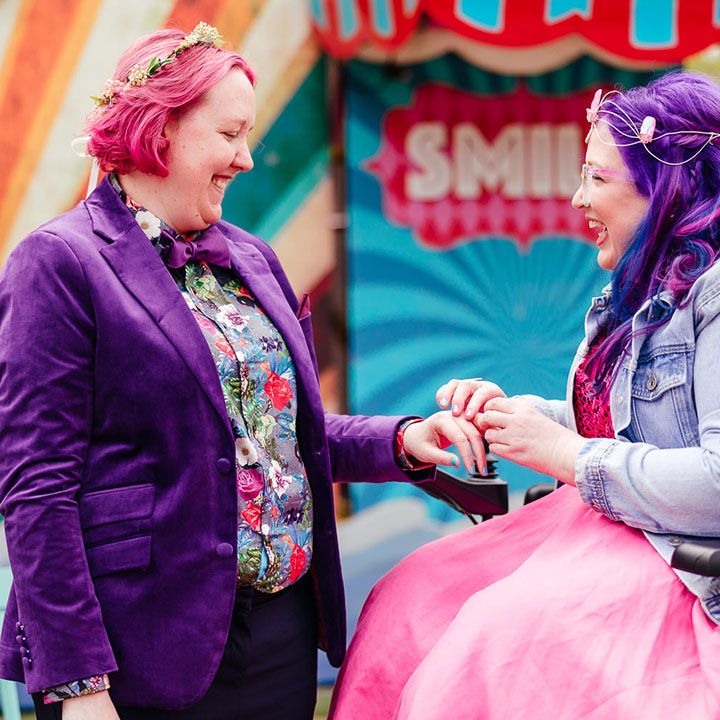 Bride in purple velvet blazer and black suit trousers with purple bowtie, pink patterned pocket square and colourful patterned shirt and bridal flower crown with bride in wheelchair wearing personalised denim jacket, pink wedding dress and gemstone bridal crown 