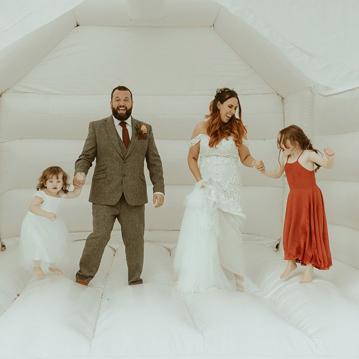 Groom in grey tweed suit with burgundy tie holding hands with daughter in white tulle dress with the bride in boho fitted lace wedding dress holding hands with daughter in burgundy dress for bouncy castle wedding 