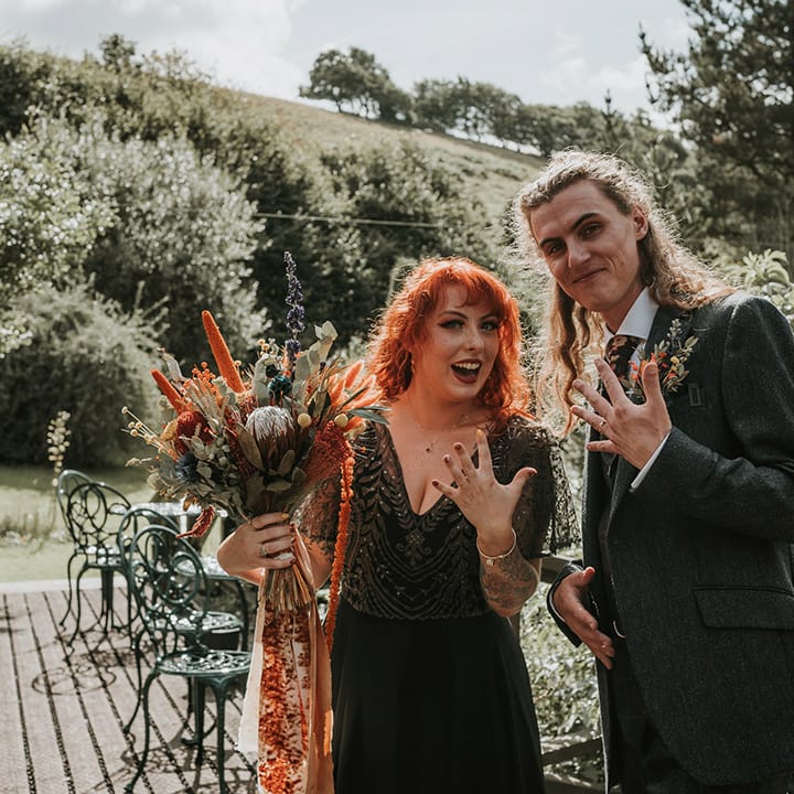Bride in detailed, embellished v neck black dress for wedding in Devon, standing with groom in dark grey three piece suit and burgundy flower patterned tie