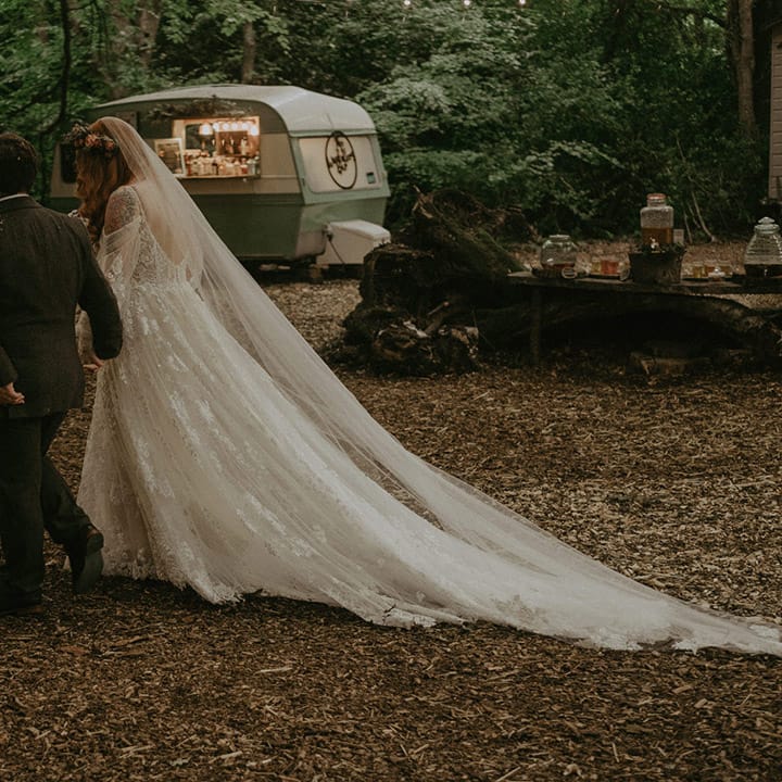 Bride in off the shoulder floral lace wedding dress walking with the groom at their woodland wedding at Lila's Wood