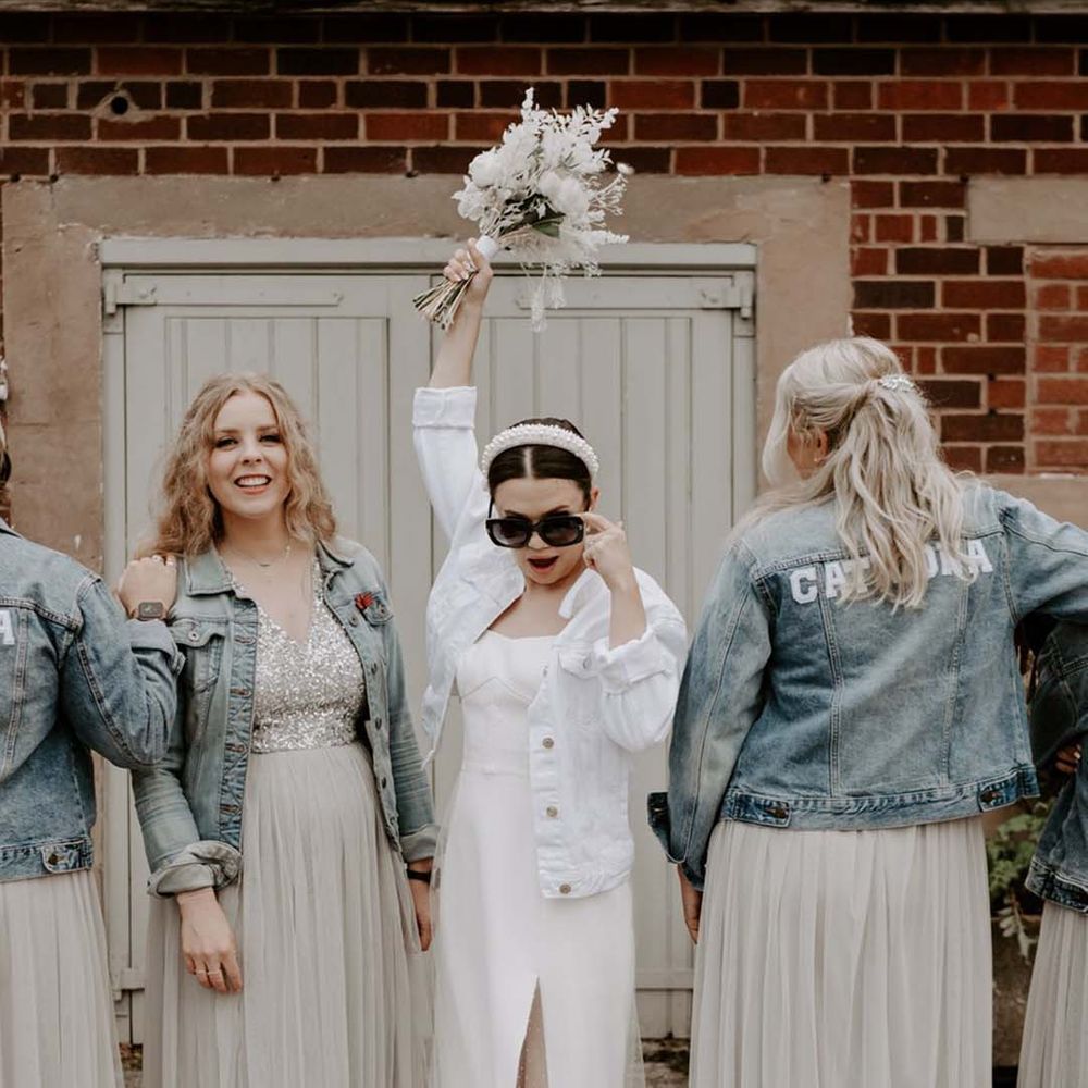 Bridesmaids in light blue denim jackets with their names on the back posing with the bride in a white bridal denim jacket and sunglasses 