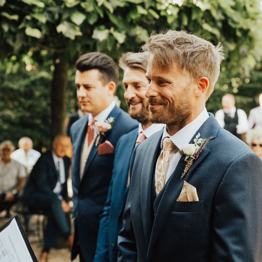 Groom in navy blue suit with gold silk tie, gold silk pocket square and rose buttonhole waits for bride