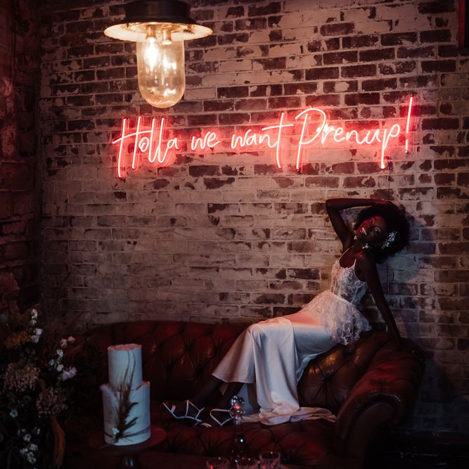 Bride in a fitted wedding dress with lace bodice and satin skirt sitting on a sofa next to a red neon wedding sign 