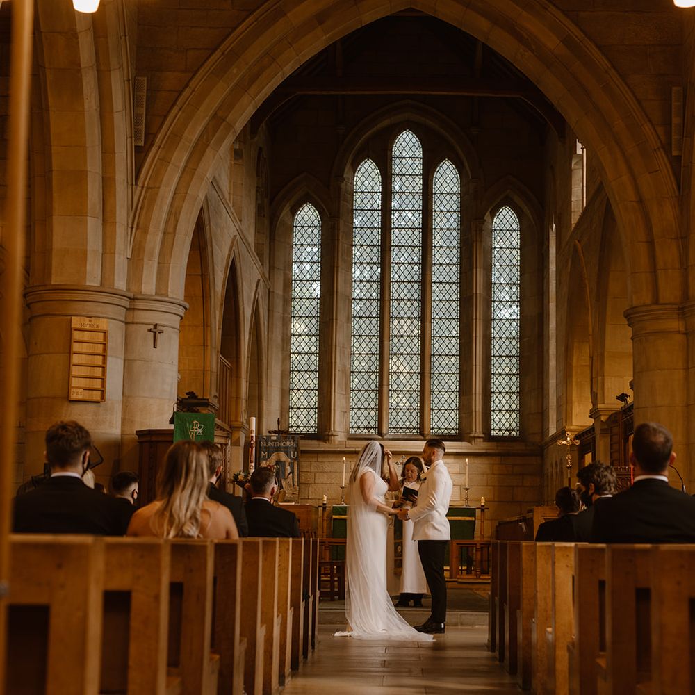 Groom in white tuxedo jacket holds hands with bride in white Made With Love wedding dress and white chapel length veil at the church altar