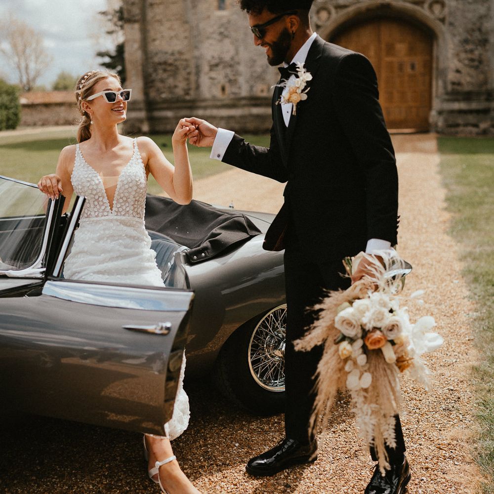 Groom in a tuxedo helping his bride in an appliqué wedding dress with deep V front out of their vintage wedding car 