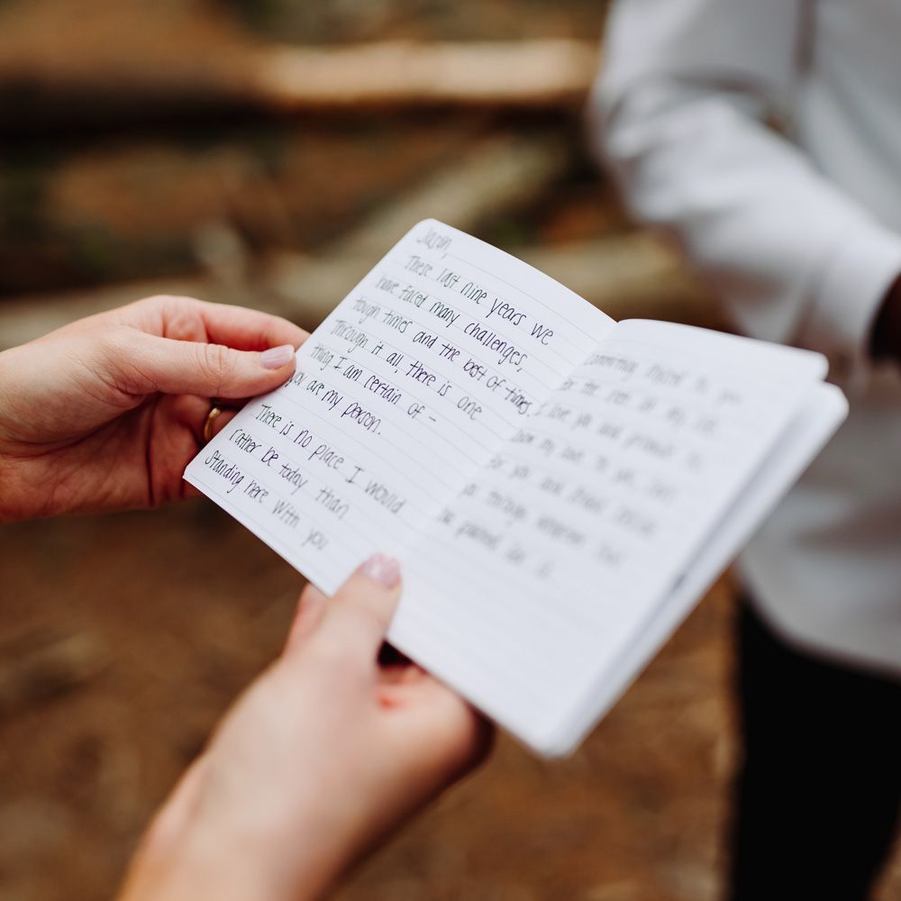 Bride holds notebook with private handwritten vows as she stands in woodland with groom in white tuxedo jacket at backyard wedding