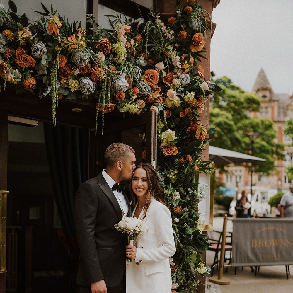 Groom in a black tuxedo kissing his bride's forehead in a doorway decorated with flowers 