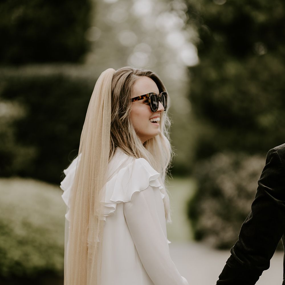 Bride in a short ruffle sleeve wedding dress wearing sunglasses and a beige coloured veil 