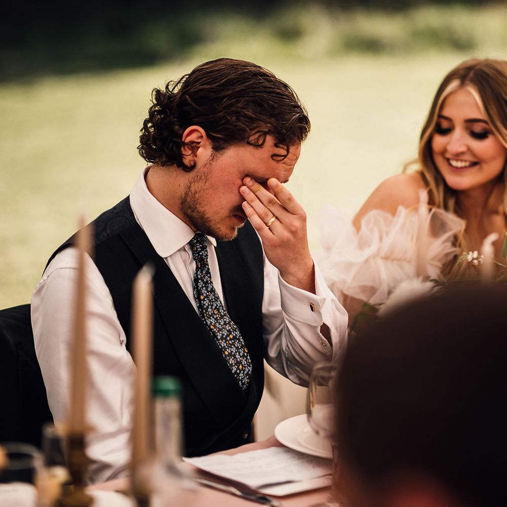 Emotional groom in white shirt, blue waistcoat and patterned tie holds hands to eyes as bride in Halfpenny London Mayfair dress smiles during marquee wedding reception in Cornwall