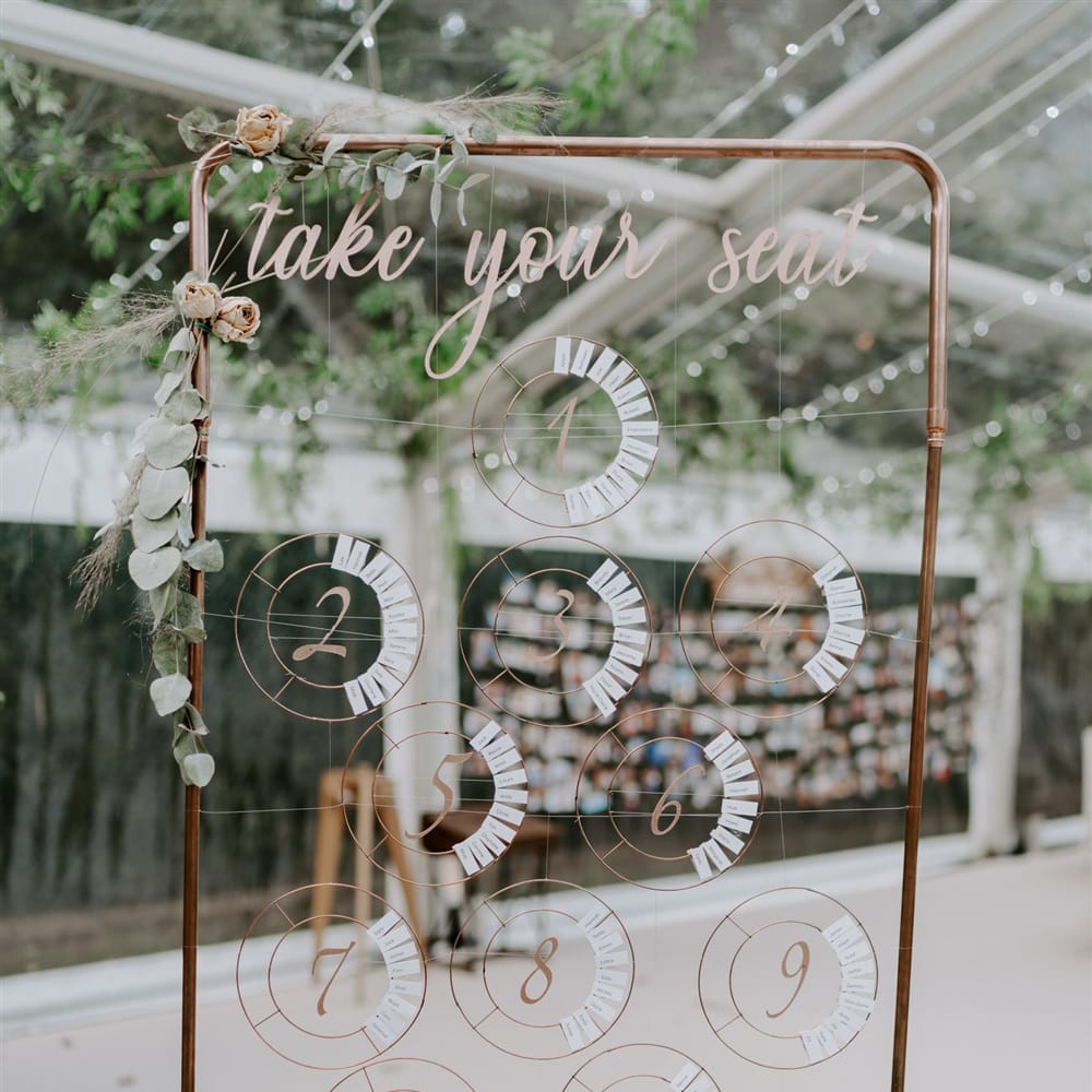 Copper frame seating plan with circular numbers and names strung across and foliage decor for marquee wedding reception