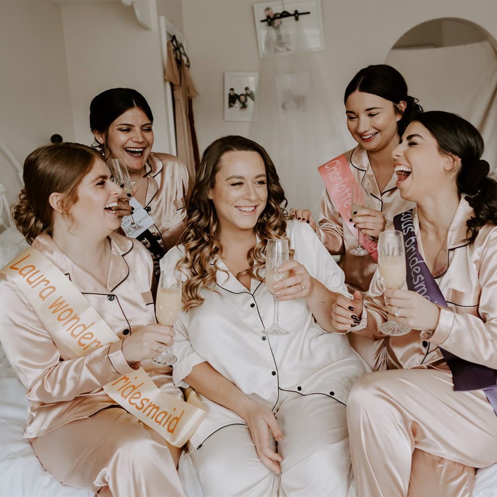 Bride in white satin pyjamas sits on double bed with bridesmaids in ivory satin pyjamas and sashes as they all hold glasses of sparkling wine before DIY garden wedding in Bedfordshire
