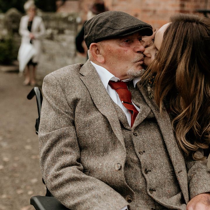 Female wedding guest in a brown satin dress kissing a male guest in a beige wool suit and flat cap