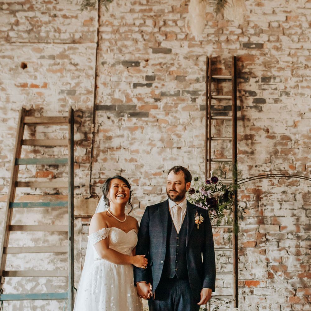 Bride in lace wedding dress and pearl necklace laughs whilst holding arm of groom in blue check three piece suit and light pink tie during wedding ceremony