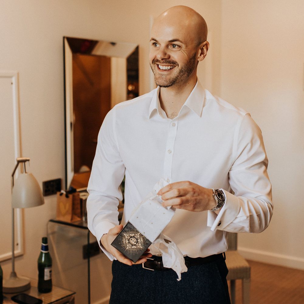 Groom in white shirt and dark trousers opens gift before wedding