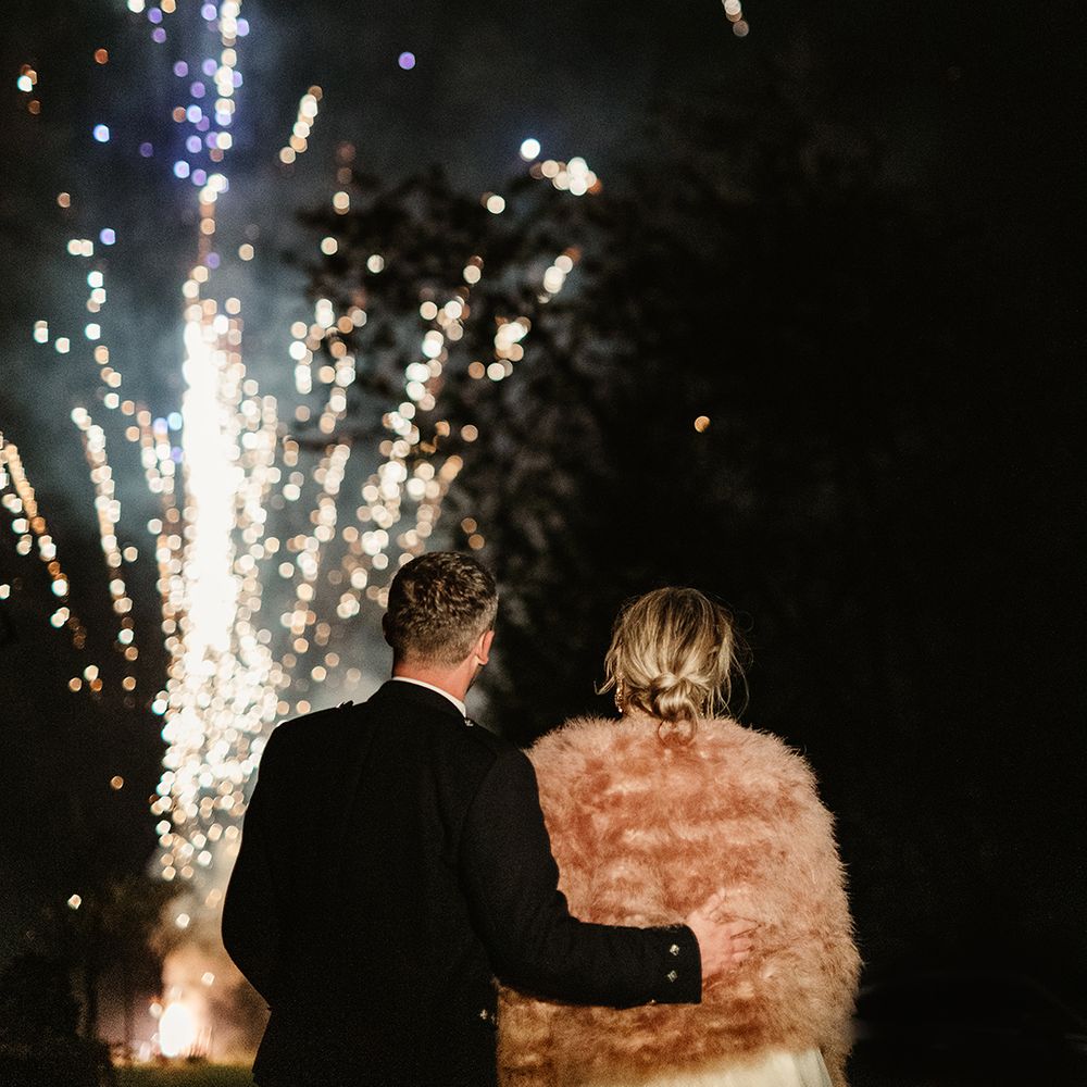 Bride in pink fur jacket and groom in black tie and kilt stands with arm around bride as they watch fireworks at the end of their wedding day