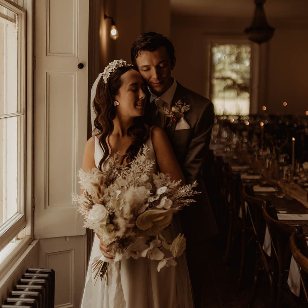 Bride with white flower earrings and matching headband in Alexandra Grecco wedding dress holding white flower and dried grass bouquet with groom in neutral suit with dried flower buttonhole