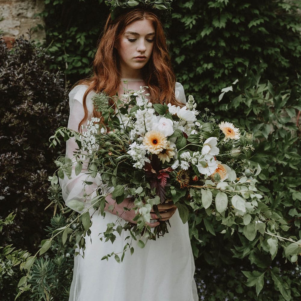 Bride in a sheer long sleeve wedding dress holding an oversized green foliage, white and yellow flower wedding bouquet 