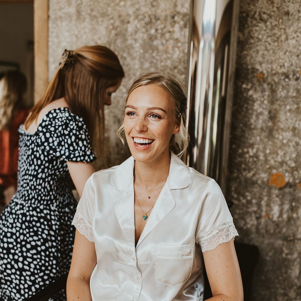 Bride smiles wearing white satin pyjamas with lace cuff detail as she gets ready for her wedding day 