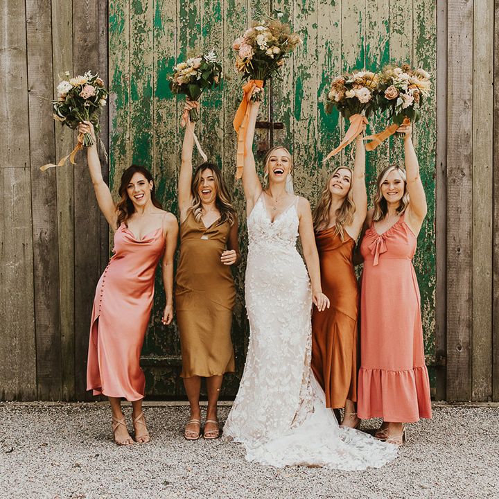 Bride in lace Made with Love wedding dress and bridesmaids in mismatched dresses lifting their bouquets in the air 