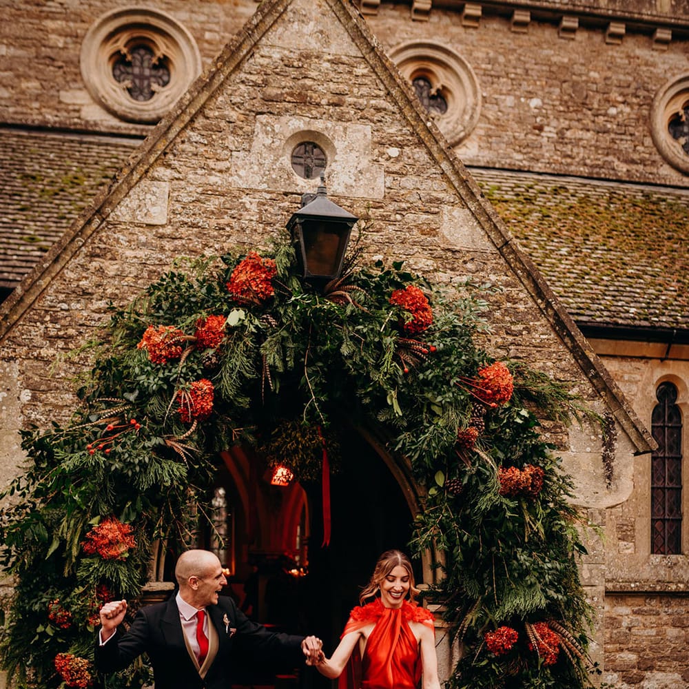 Bride in satin red wedding dress with cape and groom in three piece suit with red tie holding hands outside large foliage and red flower wedding arch