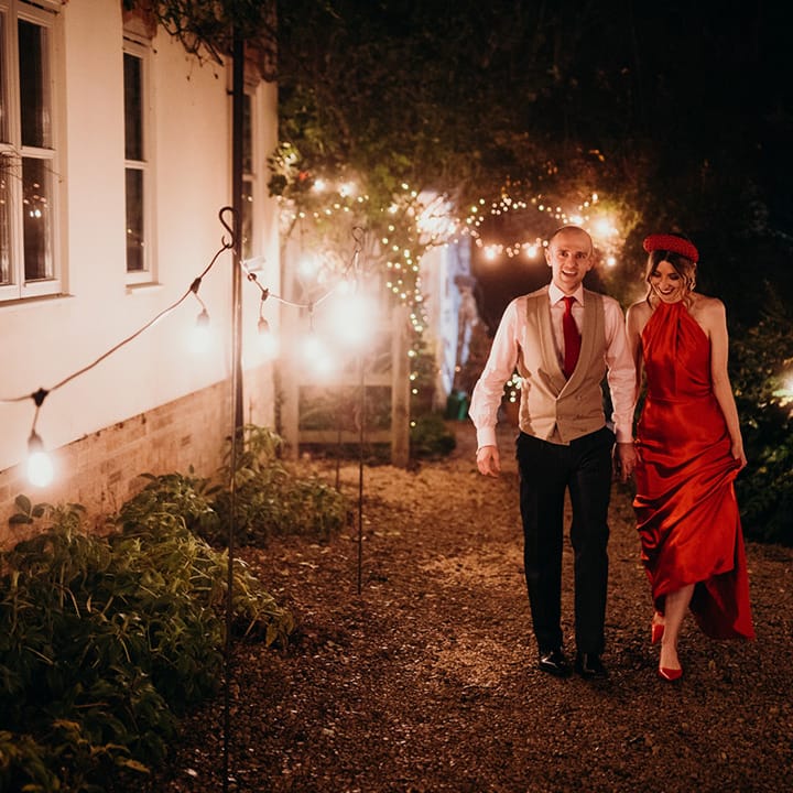 Bride in red silk wedding dress with cape and large embellished red headband and groom in three piece suit with red tie holding hands surrounded by fairylights at winter wedding