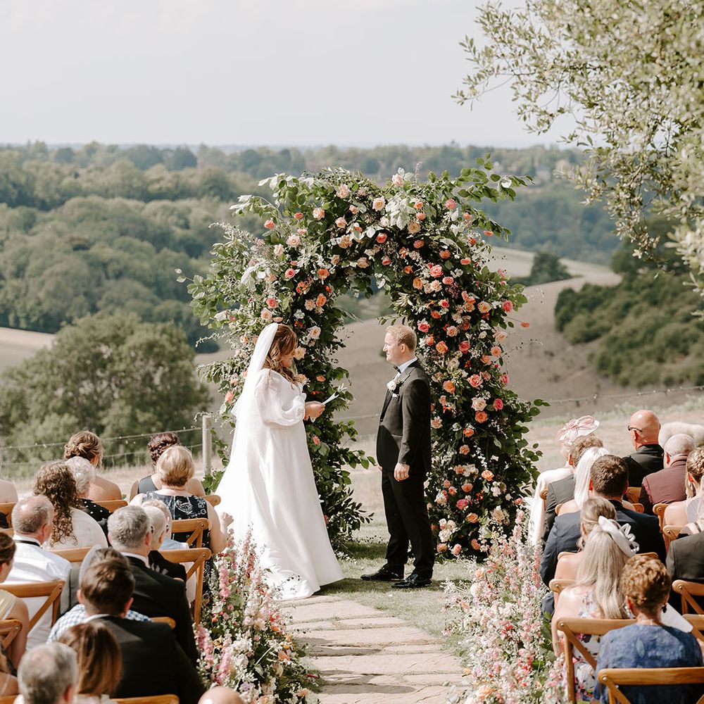 Bride & groom stand in front of beautiful floral archway with pops of orange during outdoor wedding ceremony