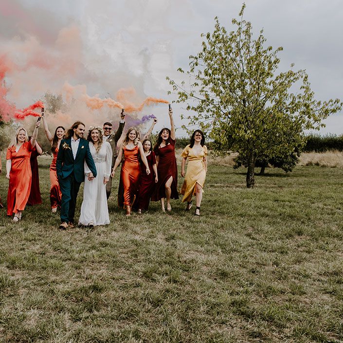Bride & groom walk with wedding party in Autumnal bridesmaid dresses whilst holding colourful smoke bombs 